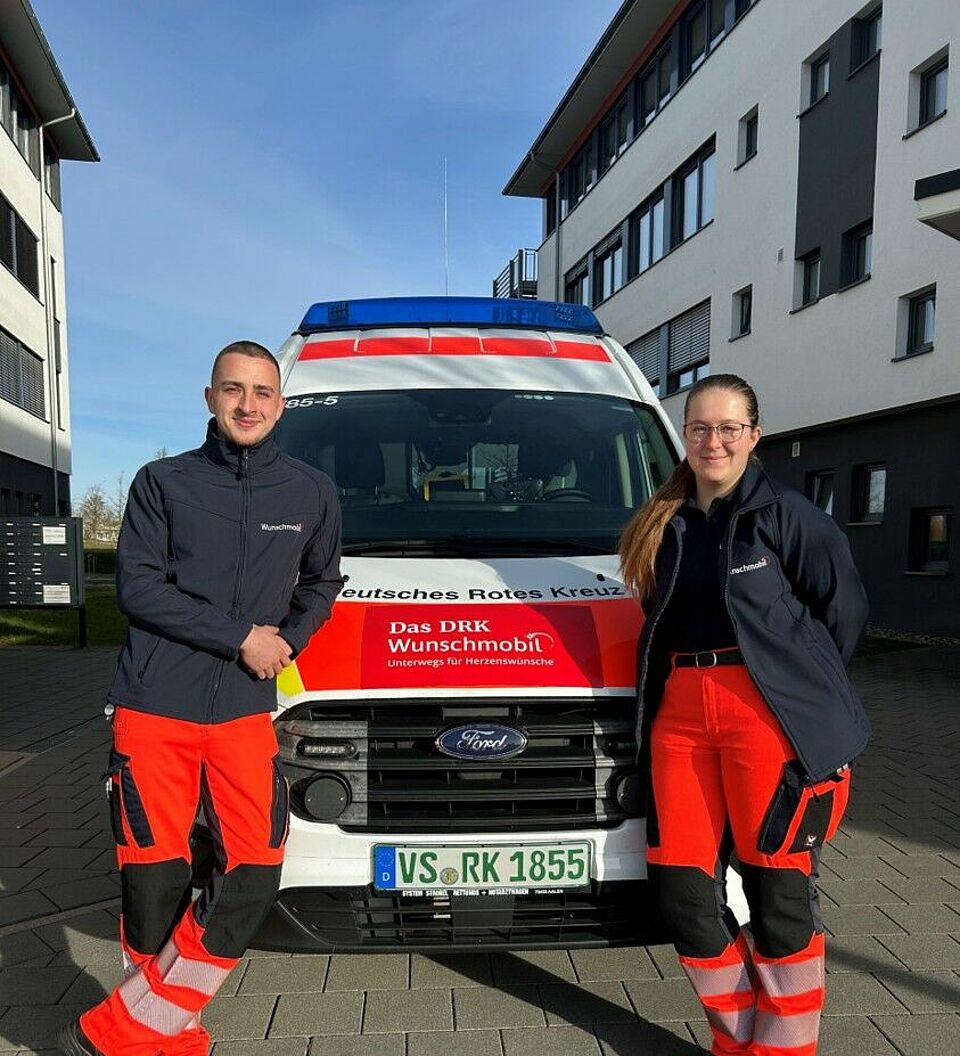 RS Ein junger Mann und eine junge Frau in DRK Kleidung lehnen rechts und links an der Motorhaube eines KTW