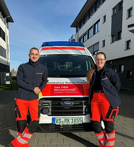 Ein junger Mann und eine junge Frau in DRK Kleidung lehnen rechts und links an der Motorhaube eines KTW