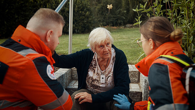 Eine alte Frau sitzt auf den Treppenstufen in Ihrem Garten. Sie trägt den Hausnotrufsender um den Hals. Vor ihr sitzen zwei Rettungssanitäter von hinten, die mit ihr sprechen.